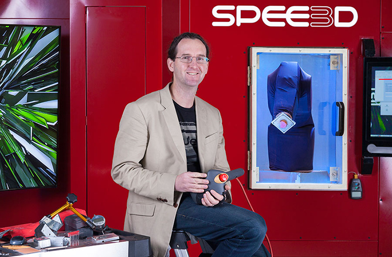 Person sitting from of large metal box machine the size of a small room. It has a window in one side showing a fabric covered robotic arm.