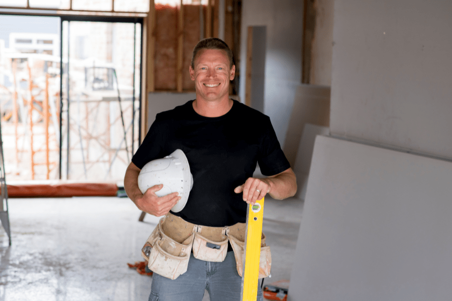 A male builder is standing in a room that's under construction. He is wearing a tool belt and holding a hard hat and spirit level.